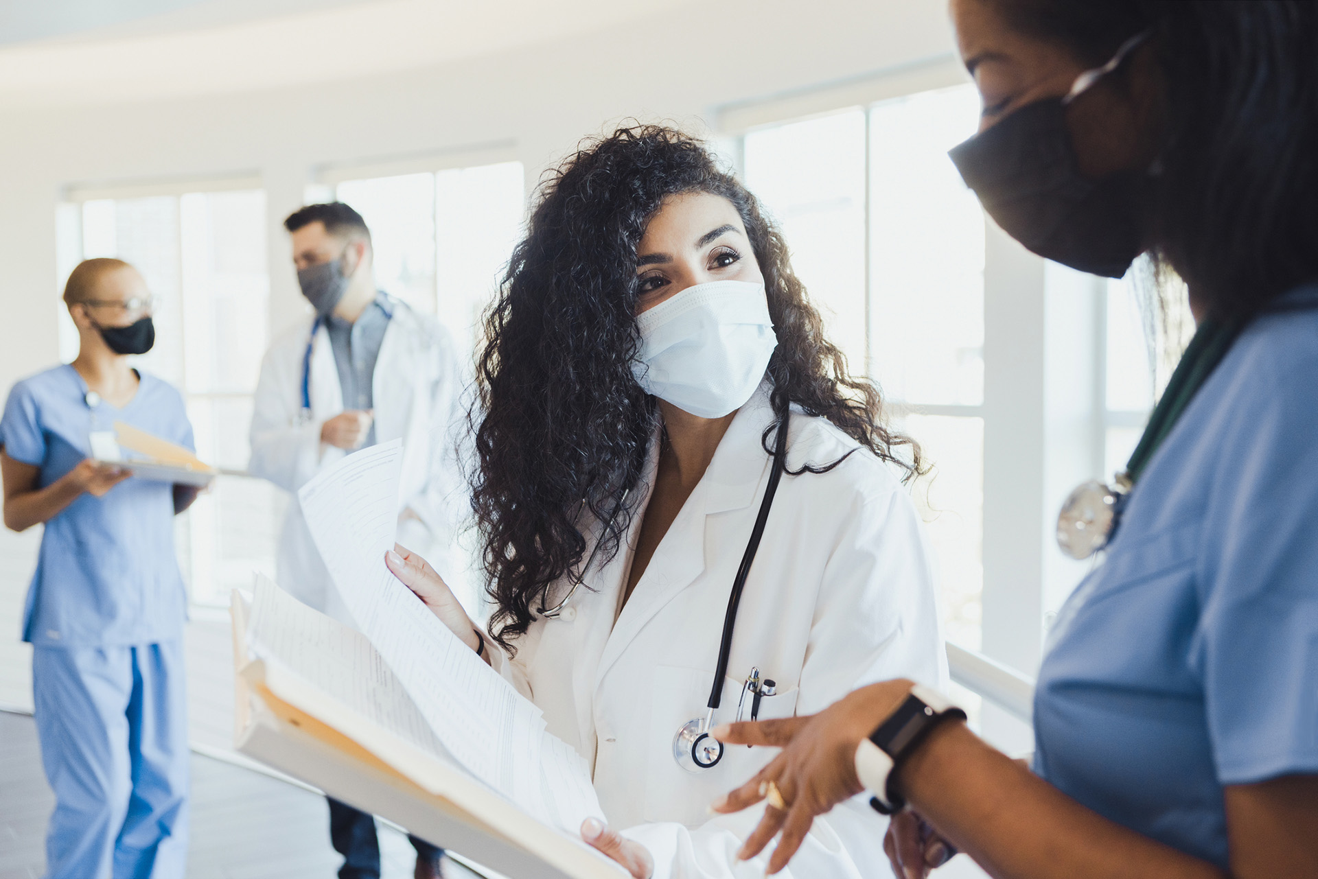 Female doctors discuss patient records in the foreground, while in the background a mid adult male doctor answers a female nurse's questions.  Everyone wears protective masks.